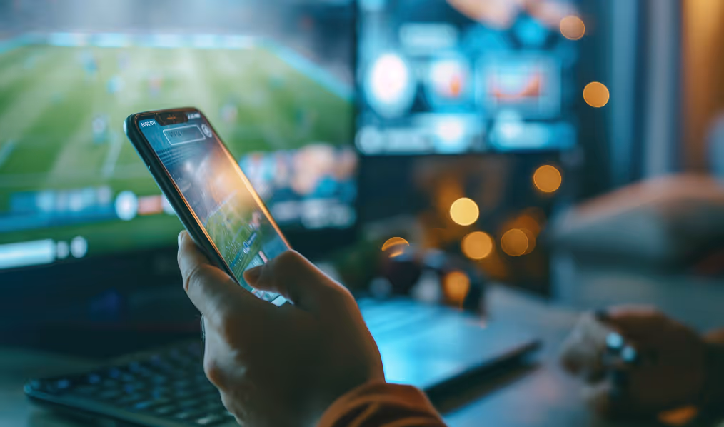 Man holding smartphone with iGaming sports betting app while watching football match on a computer screen