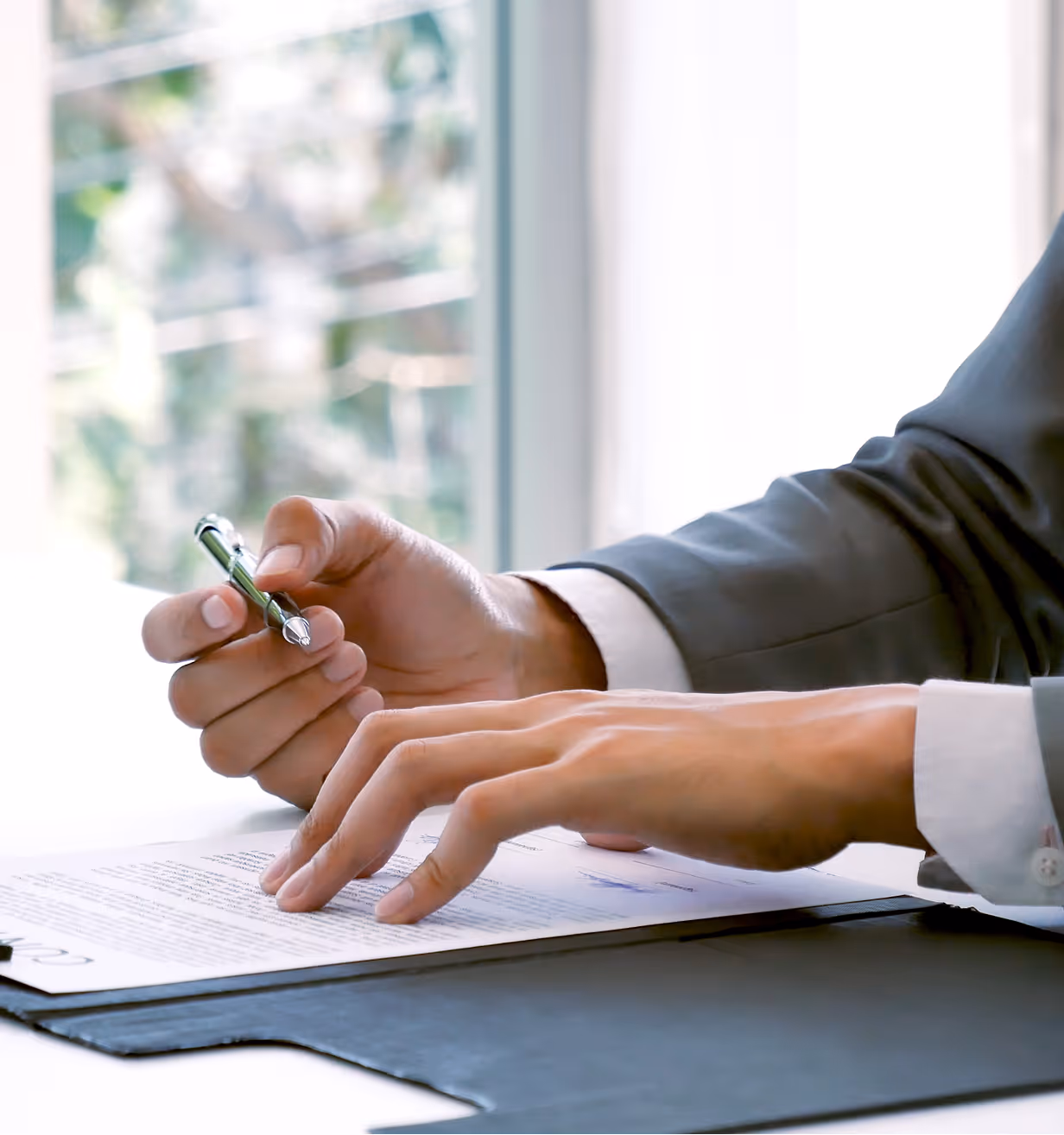 Person in a suit reviewing and holding a pen over a signed document on a desk with natural light from a window.