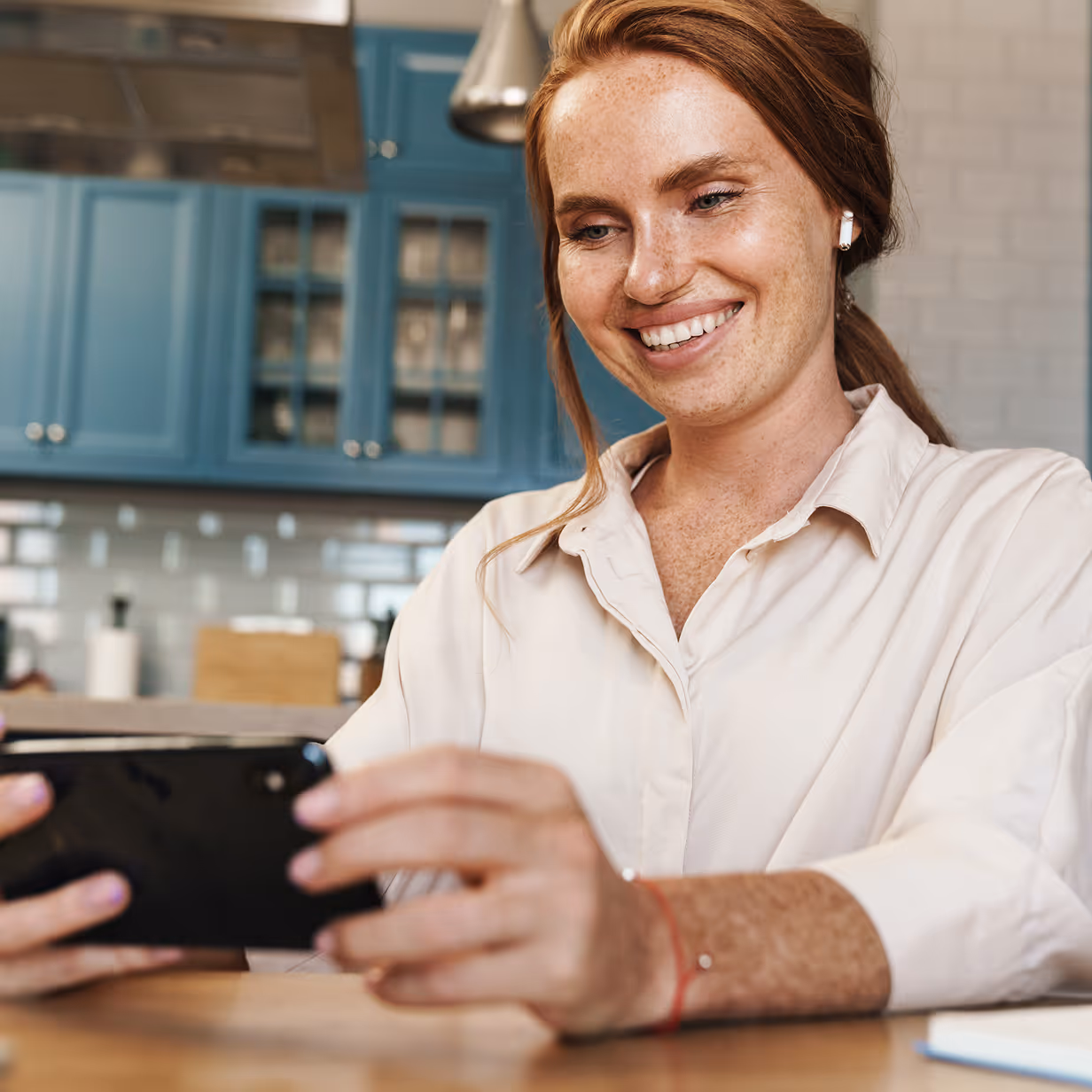 Smiling woman with red hair using a smartphone at a kitchen table.