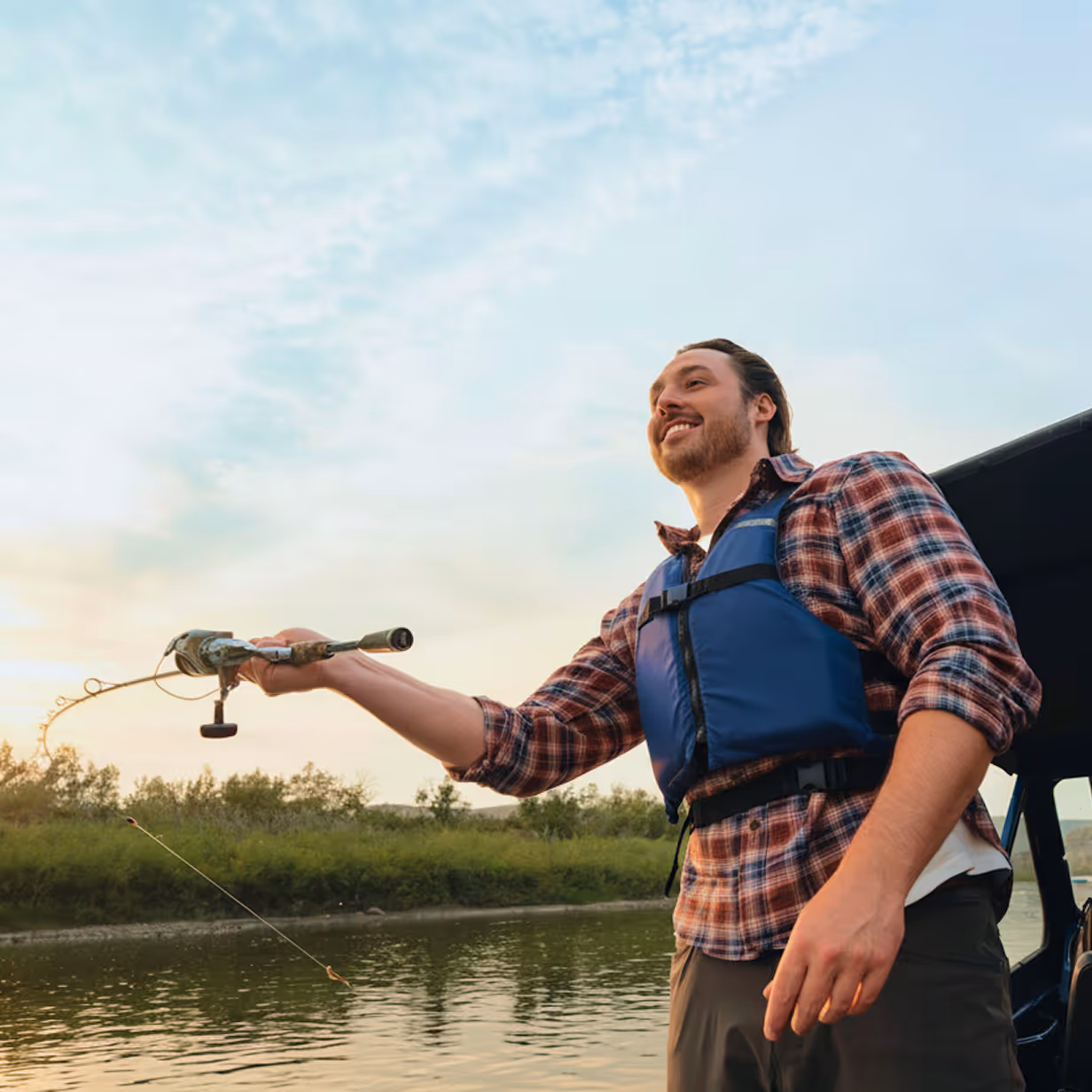 Smiling man wearing a blue life jacket and plaid shirt fishing from a boat on a calm river at sunset.