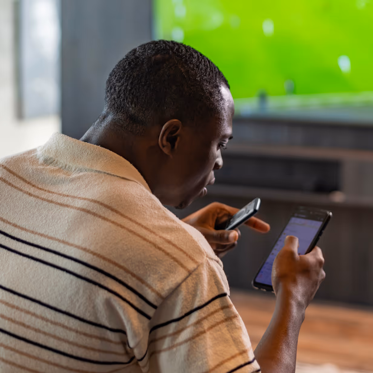 Man holding smartphone with iGaming sports betting app while watching football match at home in living room.