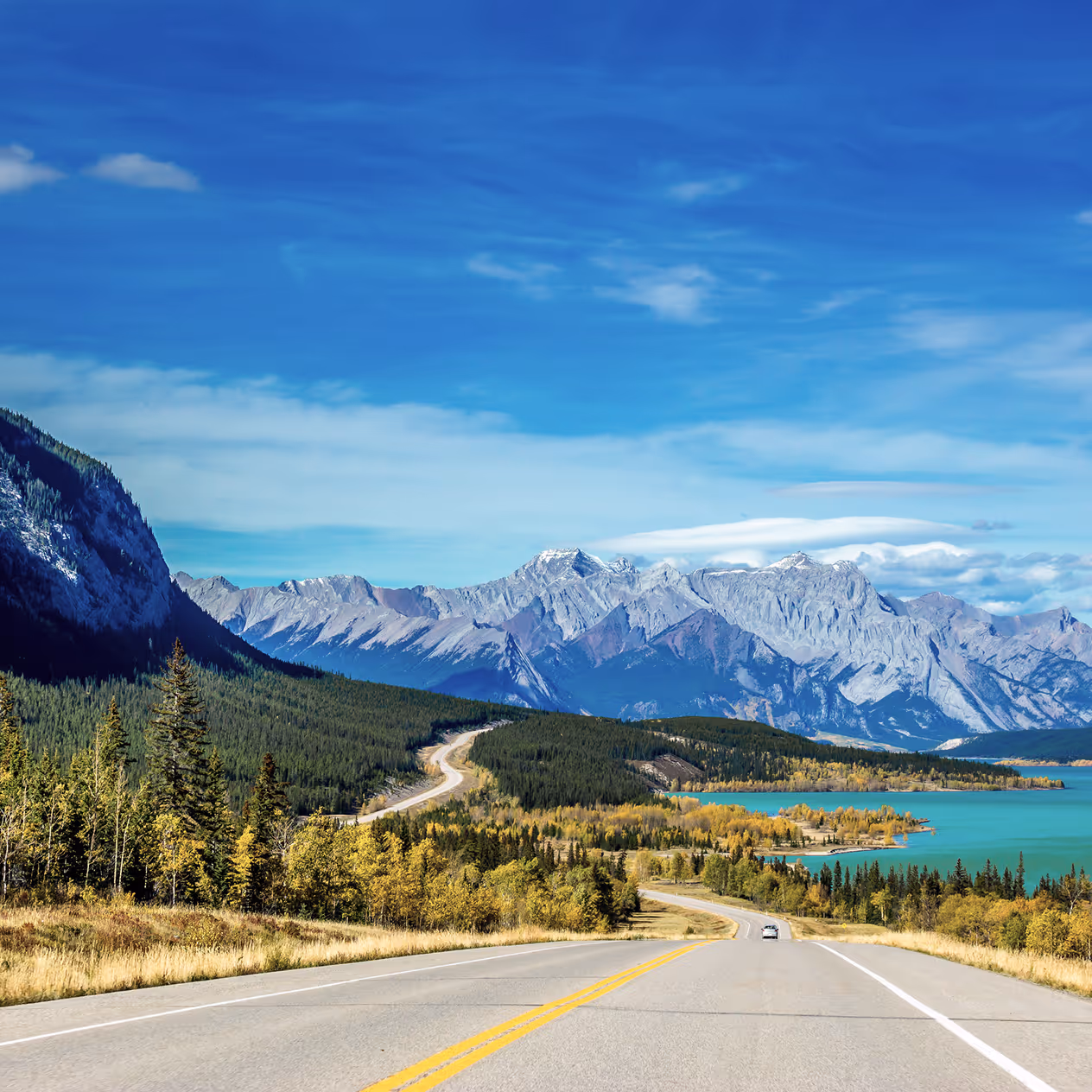 Two-lane road leading through a forested landscape with a turquoise lake on the right and snow-capped mountains under a blue sky.