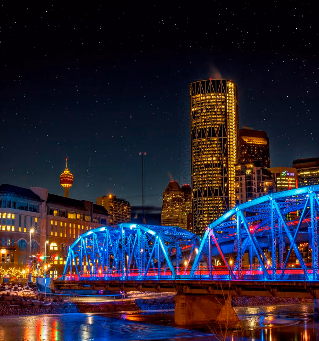 Night view of a city skyline with illuminated buildings and a blue-lit bridge over a river reflecting the lights.