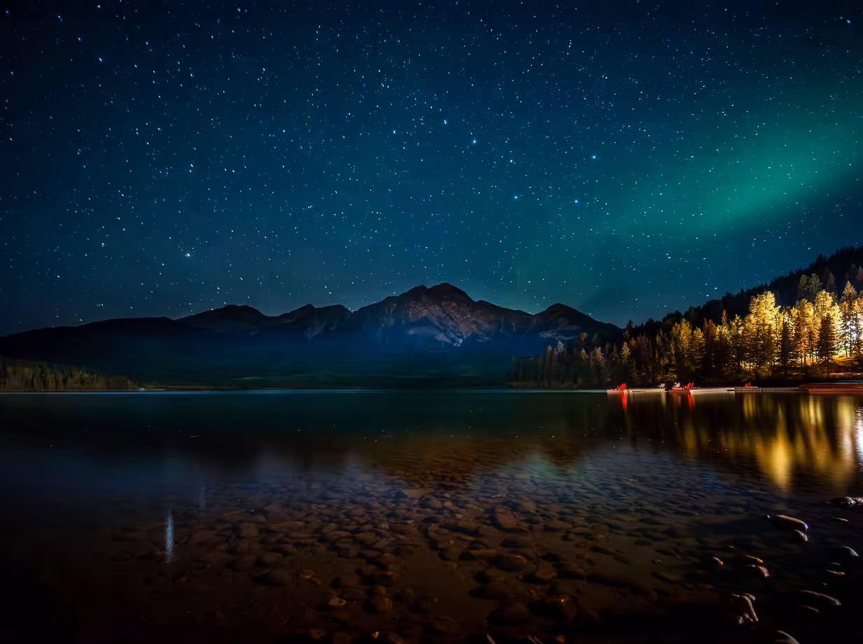 Night sky full of stars above a mountain range reflected in a calm lake with illuminated trees on the right shore.