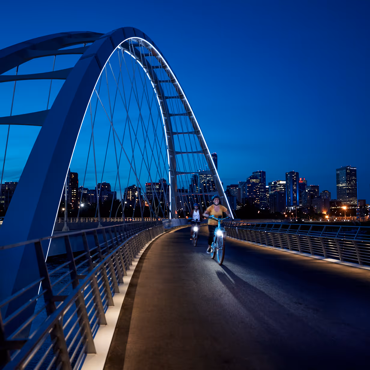 Two cyclists riding on an illuminated modern bridge at dusk with city skyline in the background.